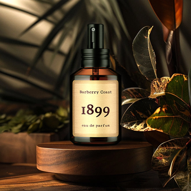 1899 Eau de Parfum Cologne by Barberry Coast. Standard 2oz amber bottle with spray cap sitting atop a circular walnut podium with tropical foliage and British Colonial decor in the background. Fragrance inspired by Ernest Hemingway and his year of birth.