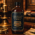 A bottle of Barberry Coast American Reserve 250 Aftershave Splash stands on a wooden table in a vintage, warm-toned room setting with a brass lamp, books, and a compass.