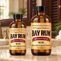 Barberry Coast Bay Rum No. 28 Aftershave Splash in 4 oz and larger 8 oz amber bottles with vintage yellow labels, side by side on a cream marble countertop in a British Colonial bathroom with dark wood, brass fixtures and tropical greenery.