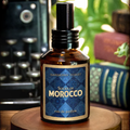 Souks of Morocco Cologne on a wood surface with a typewriter and books in the background - the glass bottle is a clear amber colored apothecary style cologne bottle with a black atomizer spray cap. Glass bottle has a blue and gold label with the words "Souks of Morocco eau de parfum by Barberry Coast"